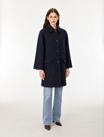 A woman modeling a dark navy blue wool-blend coat with front buttons, paired with light blue jeans and brown shoes, standing against a plain white studio background.