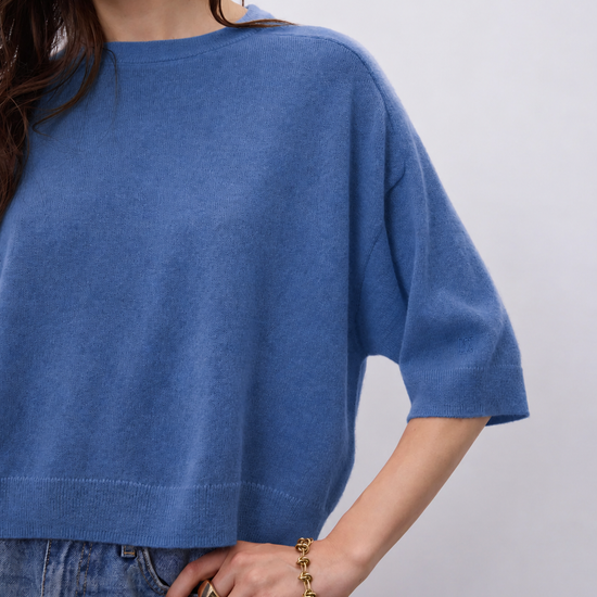 A close-up of a woman wearing a blue knit short-sleeve top with relaxed fit, paired with blue jeans, against a light gray background.