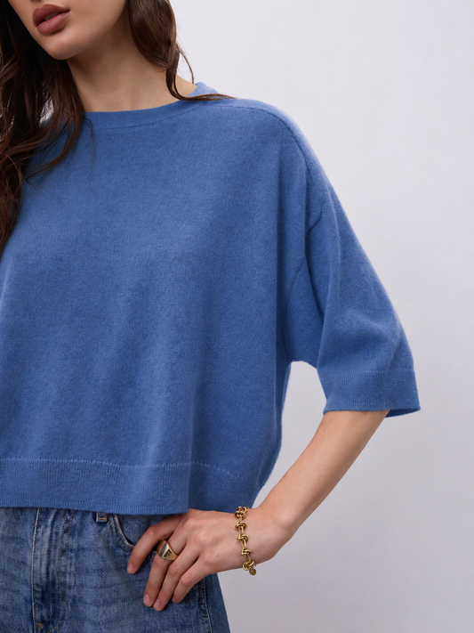 A close-up of a woman wearing a blue knit short-sleeve top with relaxed fit, paired with blue jeans, against a light gray background.