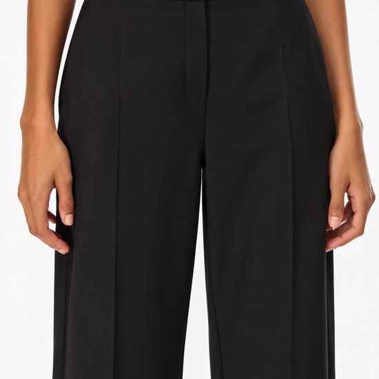 A close-up of a model wearing black wide-leg trousers with a high waist and front crease, paired with a light-colored top against a plain white background.