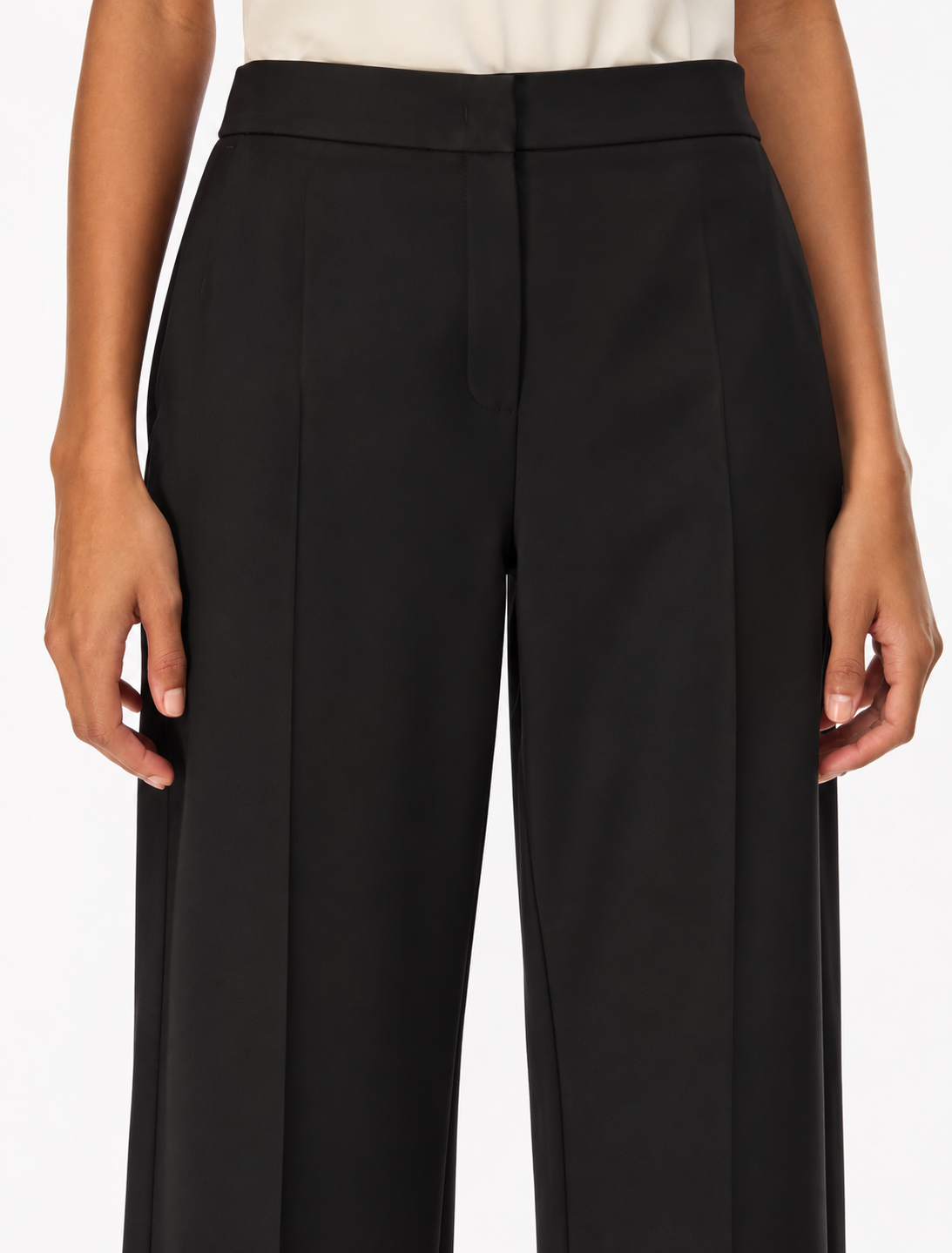 A close-up of a model wearing black wide-leg trousers with a high waist and front crease, paired with a light-colored top against a plain white background.