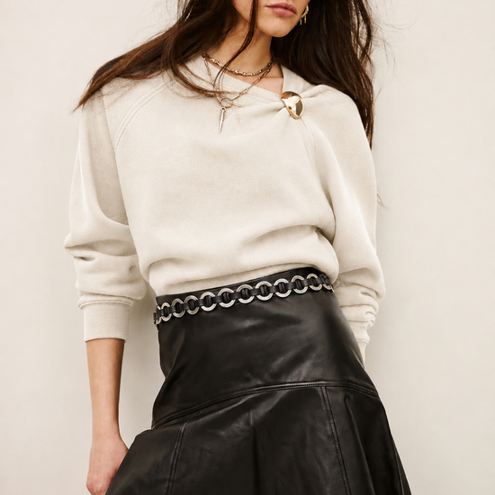 A woman modeling a cream-colored knit crewneck sweatshirt and a black faux-leather pleated mini skirt with a silver chain-link waist belt, set against a plain light beige background.