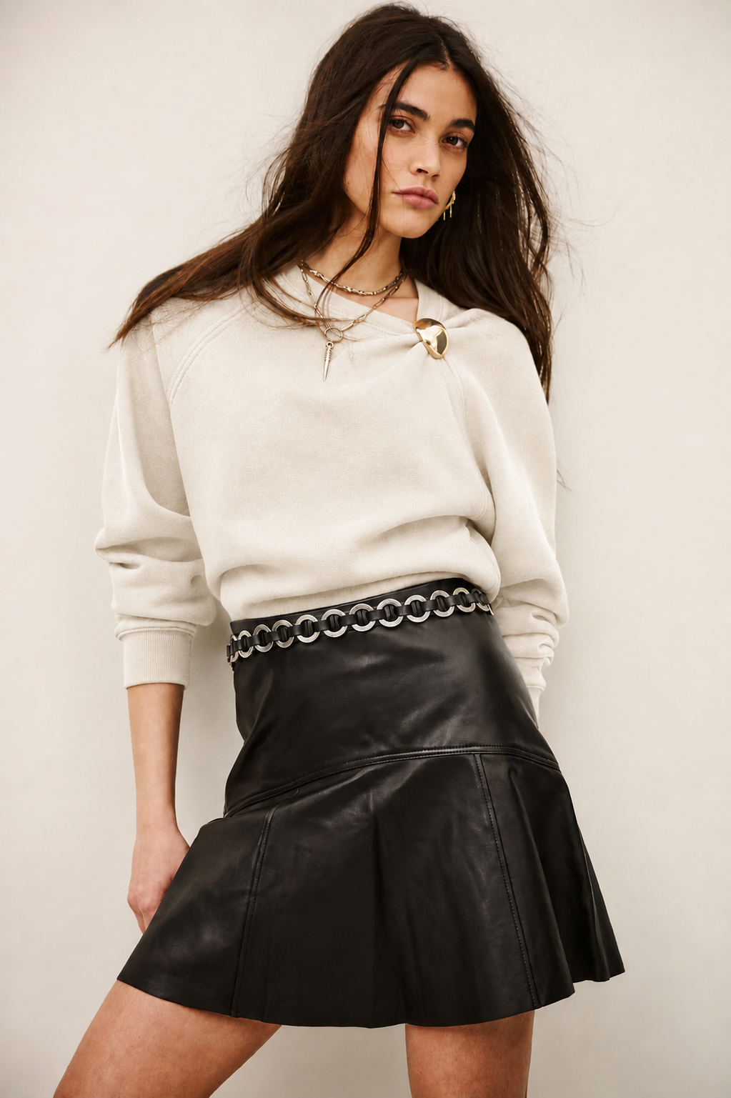A woman modeling a cream-colored knit crewneck sweatshirt and a black faux-leather pleated mini skirt with a silver chain-link waist belt, set against a plain light beige background.