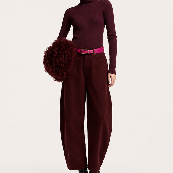 A model stands against a light gray studio background wearing a dark burgundy turtleneck sweater, matching wide-leg trousers, a bright pink belt, black pointed boots, and holding a dark burgundy fuzzy pom-pom accessory.