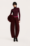 A model stands against a light gray studio background wearing a dark burgundy turtleneck sweater, matching wide-leg trousers, a bright pink belt, black pointed boots, and holding a dark burgundy fuzzy pom-pom accessory.