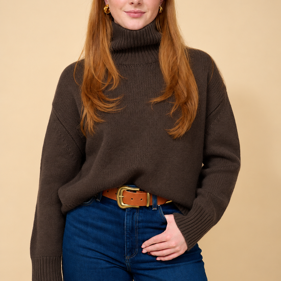 A woman with long red hair wearing a dark brown ribbed turtleneck sweater, blue jeans, and a brown belt, standing against a solid beige background.