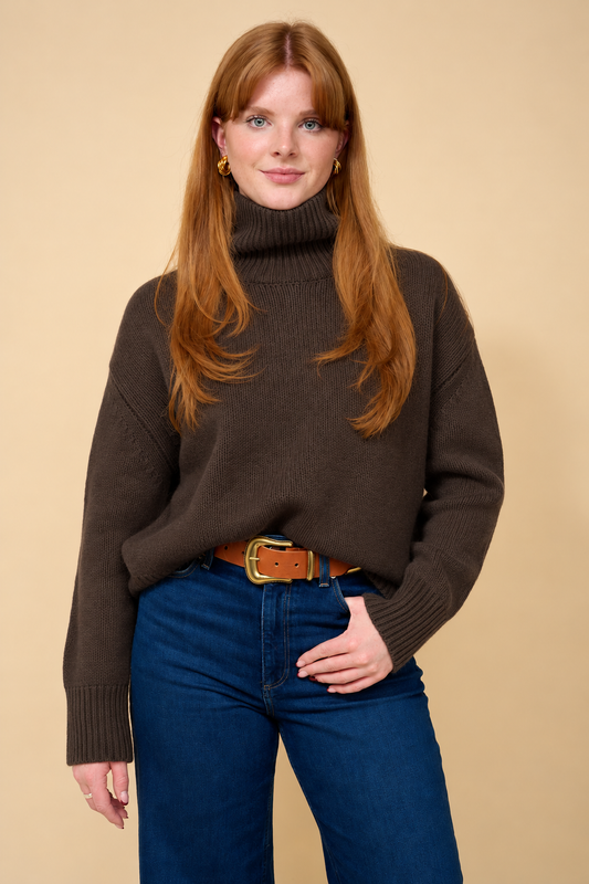 A woman with long red hair wearing a dark brown ribbed turtleneck sweater, blue jeans, and a brown belt, standing against a solid beige background.
