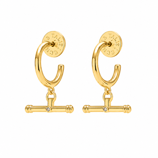 A pair of gold huggie hoop earrings with a dangling barbell charm and a small clear rhinestone in the center, shown on a plain white background.