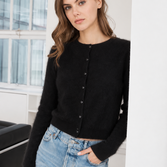 A woman wearing a black long-sleeve button-front cardigan and light blue jeans, standing in a bright indoor setting with a window and white walls in the background.