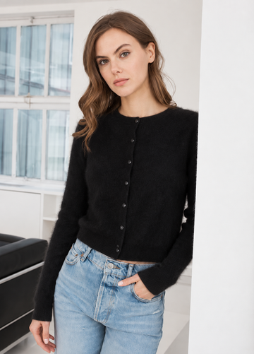 A woman wearing a black long-sleeve button-front cardigan and light blue jeans, standing in a bright indoor setting with a window and white walls in the background.