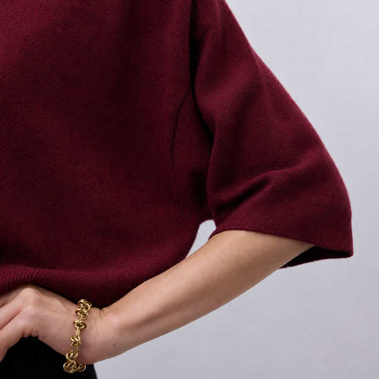Close-up of a person wearing a burgundy knit short-sleeve top with a chunky gold chain bracelet on the wrist, against a light gray background.