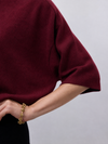 Close-up of a person wearing a burgundy knit short-sleeve top with a chunky gold chain bracelet on the wrist, against a light gray background.