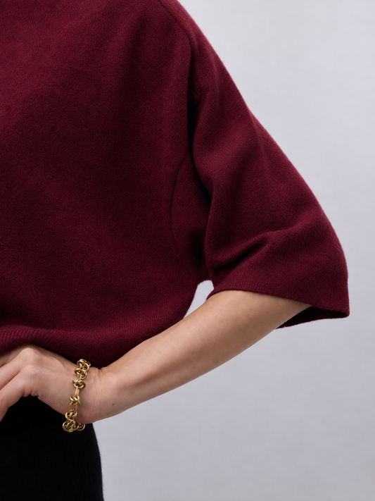Close-up of a person wearing a burgundy knit short-sleeve top with a chunky gold chain bracelet on the wrist, against a light gray background.
