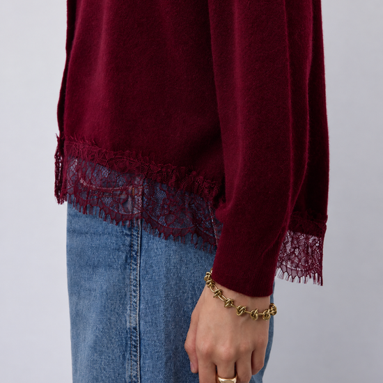 A close-up of a person wearing a long-sleeve burgundy knit top with lace trim at the hem and cuffs, paired with blue denim jeans. The person is also wearing a gold chain bracelet and ring against a plain light gray background.