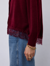 A close-up of a person wearing a long-sleeve burgundy knit top with lace trim at the hem and cuffs, paired with blue denim jeans. The person is also wearing a gold chain bracelet and ring against a plain light gray background.