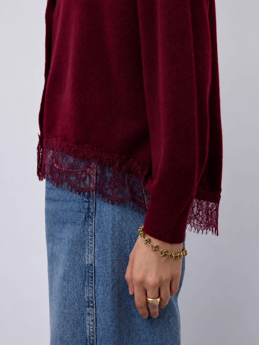 A close-up of a person wearing a long-sleeve burgundy knit top with lace trim at the hem and cuffs, paired with blue denim jeans. The person is also wearing a gold chain bracelet and ring against a plain light gray background.