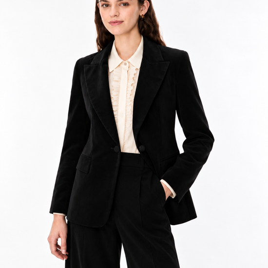 A woman modeling a black tailored blazer and matching wide-leg trousers over a cream ruffled blouse, photographed against a clean white studio background.