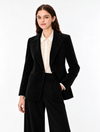 A woman modeling a black tailored blazer and matching wide-leg trousers over a cream ruffled blouse, photographed against a clean white studio background.