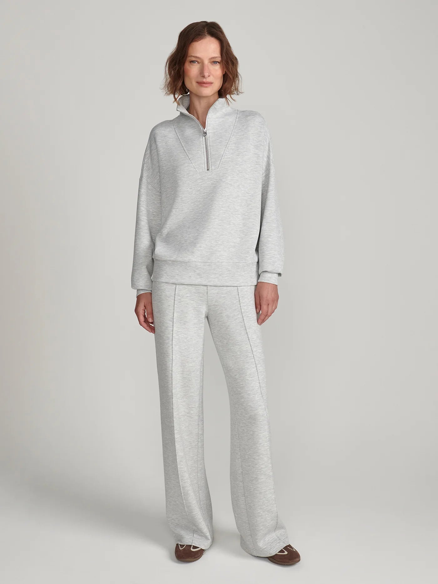 A woman modeling a light gray heathered loungewear set consisting of a quarter-zip long-sleeve top and matching wide-leg sweatpants, photographed against a neutral light gray studio background.