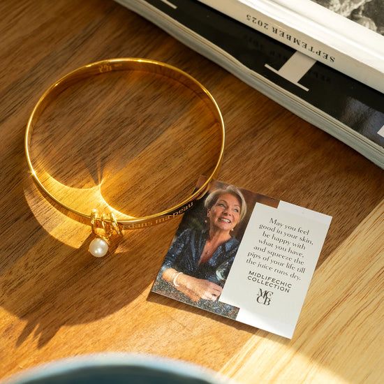 Gold bracelet with a pearl on a wooden surface next to a card featuring a woman's face and text.