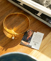 Gold bracelet with a pearl on a wooden surface next to a card featuring a woman's face and text.