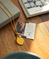 Gold necklace with a pendant on a wooden surface next to a card and book.