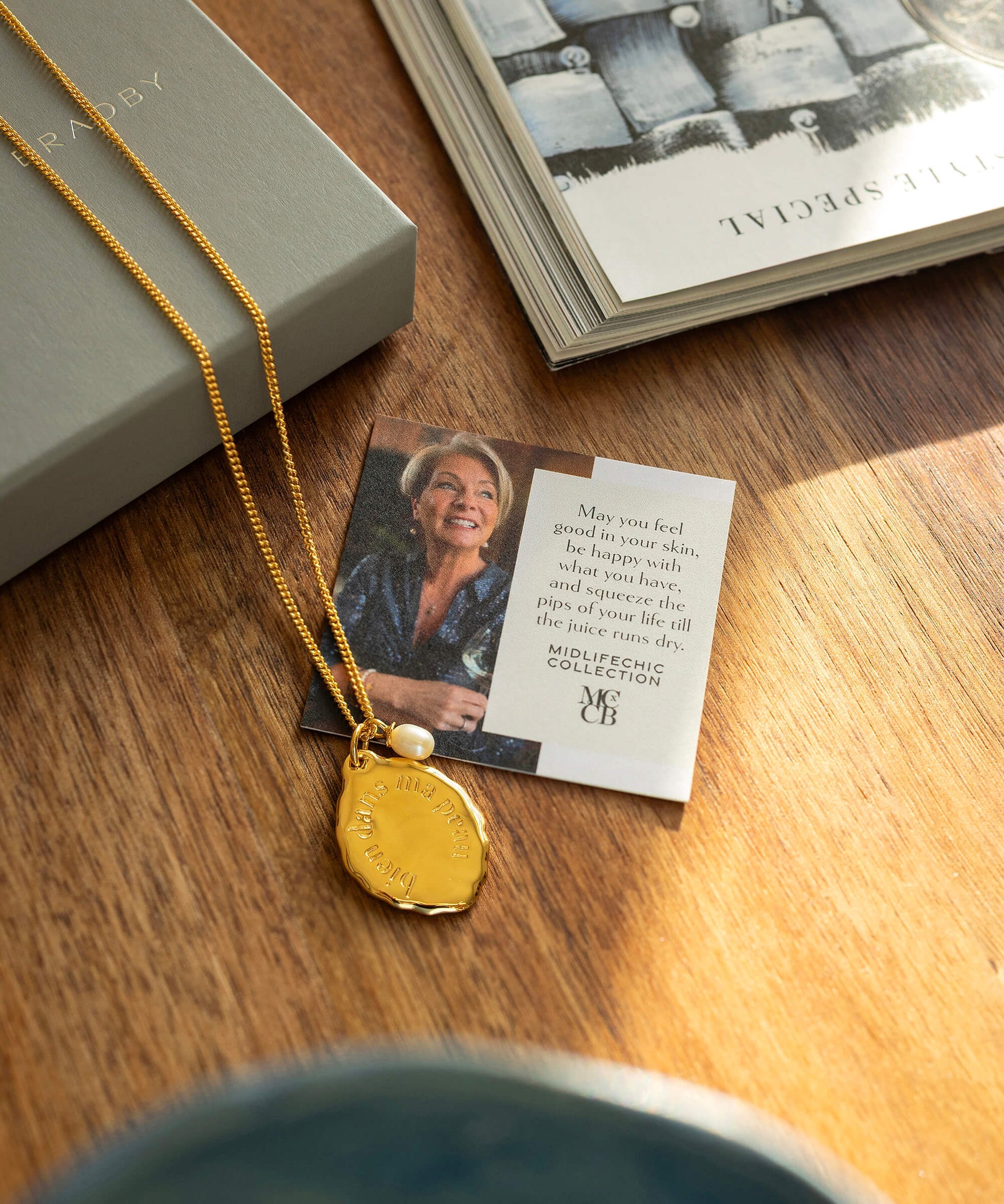 Gold necklace with a pendant on a wooden surface next to a card and book.