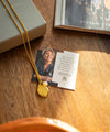 Gold necklace with a heart-shaped pendant on a wooden surface next to a card and book.