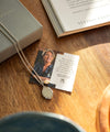 Necklace with pendant on a card featuring a photo and text, placed on a wooden surface next to an open book.