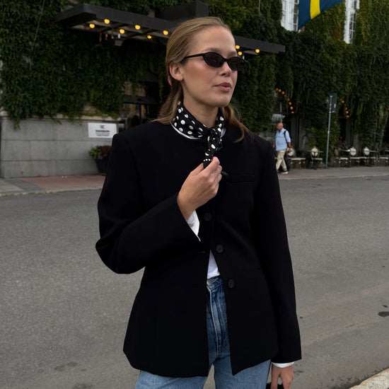 Woman in a black coat and sunglasses walking on a street with a building and flags in the background.
