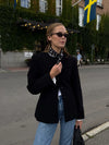 Woman in a black coat and sunglasses walking on a street with a building and flags in the background.