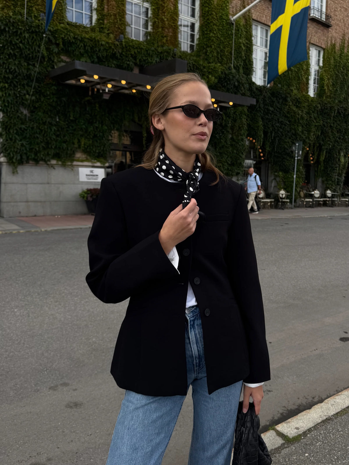 Woman in a black coat and sunglasses walking on a street with a building and flags in the background.