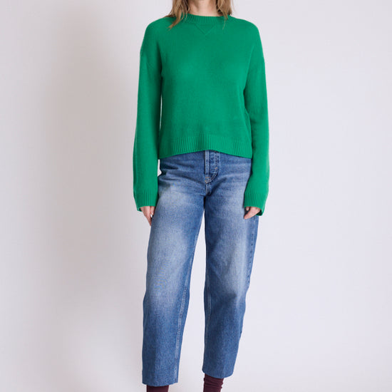 A woman modeling a bright green knit sweater, blue jeans, brown shoes, and dark socks against a plain light gray studio background.