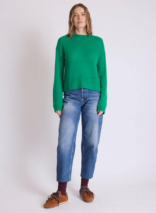 A woman modeling a bright green knit sweater, blue jeans, brown shoes, and dark socks against a plain light gray studio background.