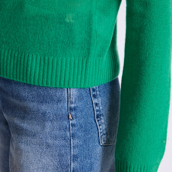 Close-up of a bright green knit sweater worn with blue jeans, showing the ribbed hem and sleeve cuff against a plain light background.