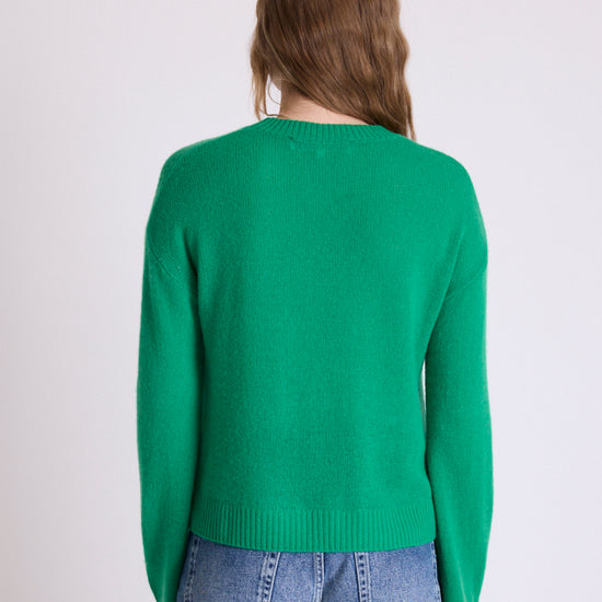 A woman viewed from the back wearing a bright green knit sweater and blue jeans against a plain light gray studio background.