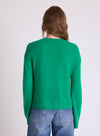 A woman viewed from the back wearing a bright green knit sweater and blue jeans against a plain light gray studio background.
