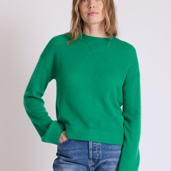 A woman wearing a bright green crewneck knit sweater and blue jeans, standing against a plain light gray studio background.