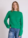A woman wearing a bright green crewneck knit sweater and blue jeans, standing against a plain light gray studio background.