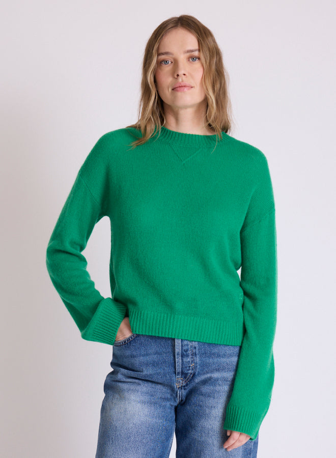A woman wearing a bright green crewneck knit sweater and blue jeans, standing against a plain light gray studio background.