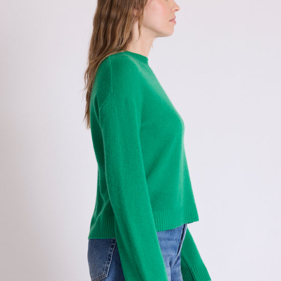 A woman shown in side profile wearing a bright green knit sweater and blue jeans against a plain light gray studio background.
