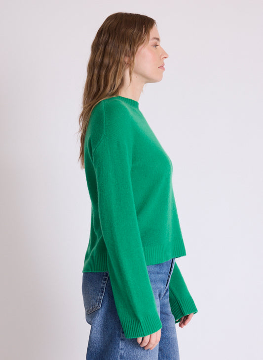 A woman shown in side profile wearing a bright green knit sweater and blue jeans against a plain light gray studio background.