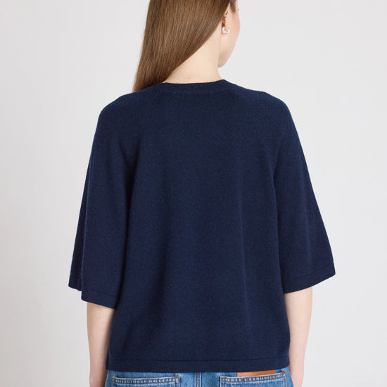 A woman viewed from the back wearing a dark navy blue short-sleeve knit top and light blue jeans against a plain light gray studio background.