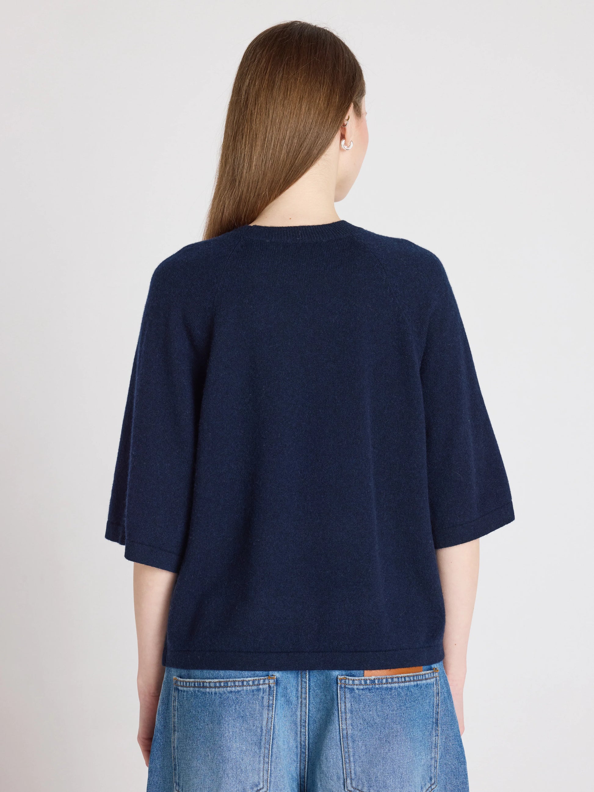 A woman viewed from the back wearing a dark navy blue short-sleeve knit top and light blue jeans against a plain light gray studio background.