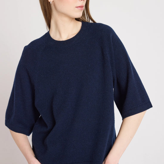 A woman wearing an oversized navy blue knit short-sleeve top and blue jeans, photographed against a plain light gray studio background.