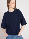 A woman wearing an oversized navy blue knit short-sleeve top and blue jeans, photographed against a plain light gray studio background.