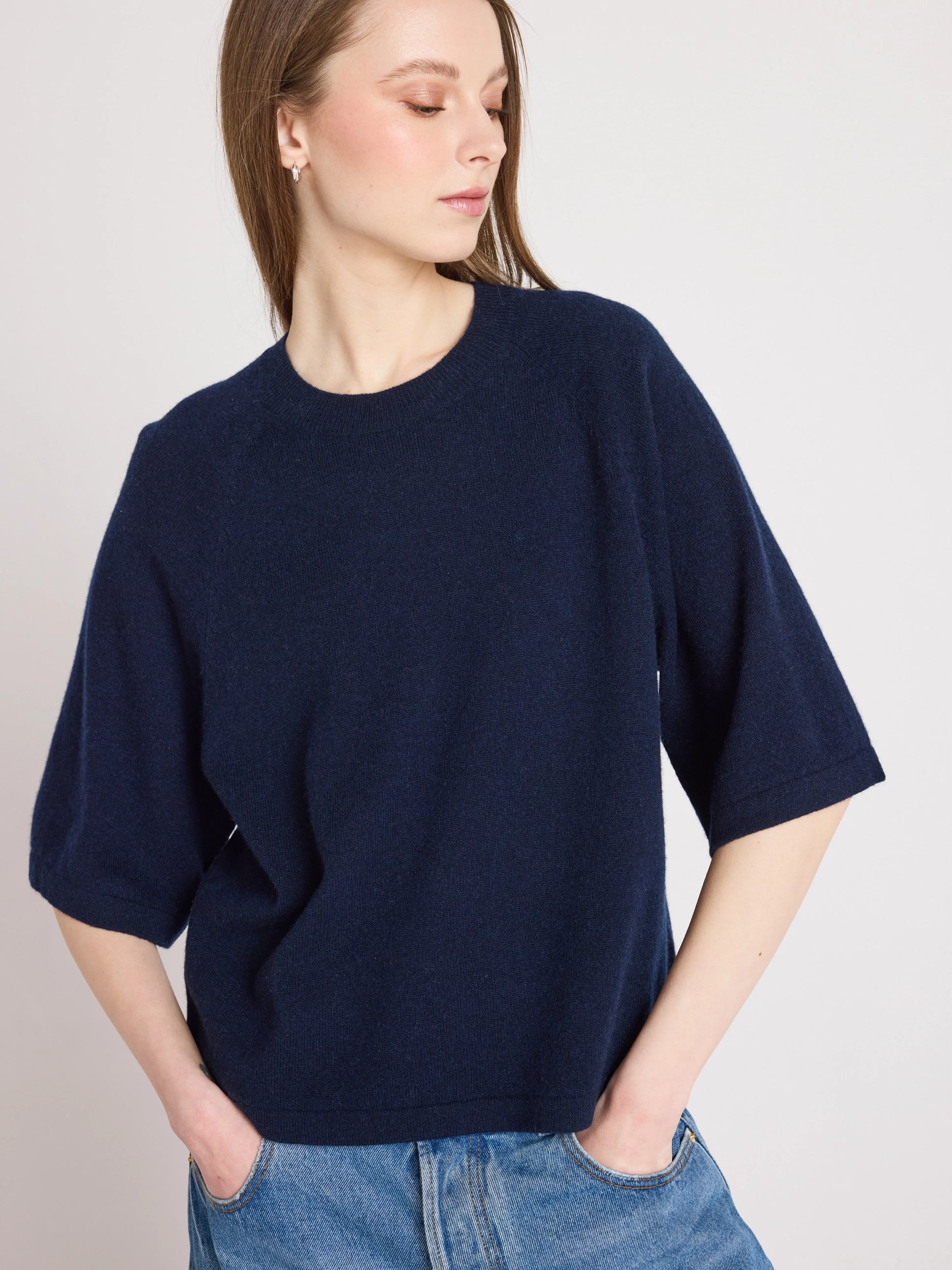 A woman wearing an oversized navy blue knit short-sleeve top and blue jeans, photographed against a plain light gray studio background.