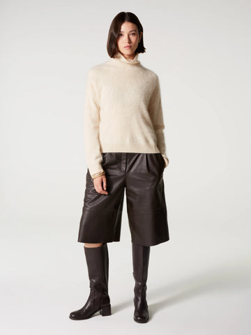 A model stands against a light gray studio background wearing a cream-colored fuzzy turtleneck sweater, dark brown leather shorts, and black knee-high boots.