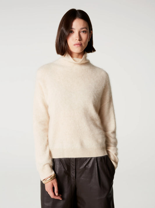 A woman with a short dark bob hairstyle wearing a fuzzy cream-colored mock neck sweater and dark brown leather pants, standing against a light gray studio background.