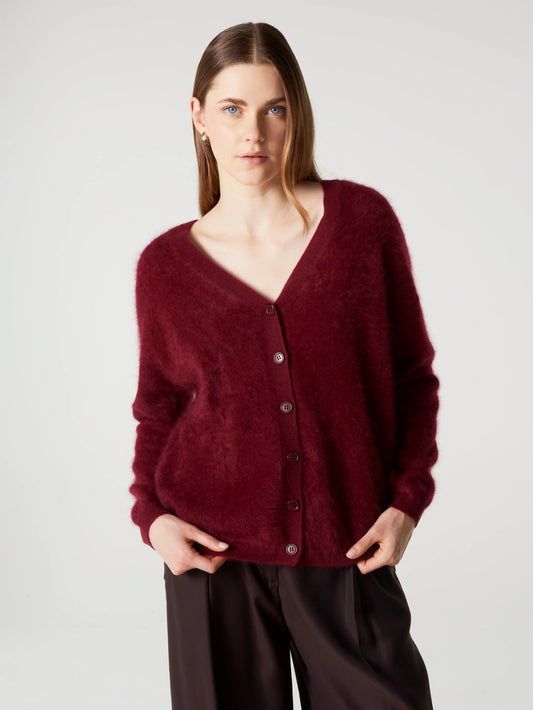 A woman wearing a fuzzy, plush button-up cardigan in deep burgundy, paired with dark brown trousers, photographed against a neutral light gray studio background.